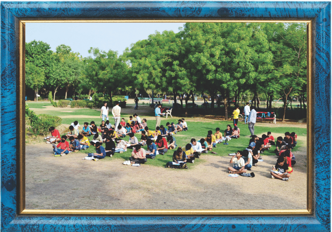 This photo shows a group of students sitting outdoors on a lawn, studying or taking an exam, surrounded by greenery and trees.