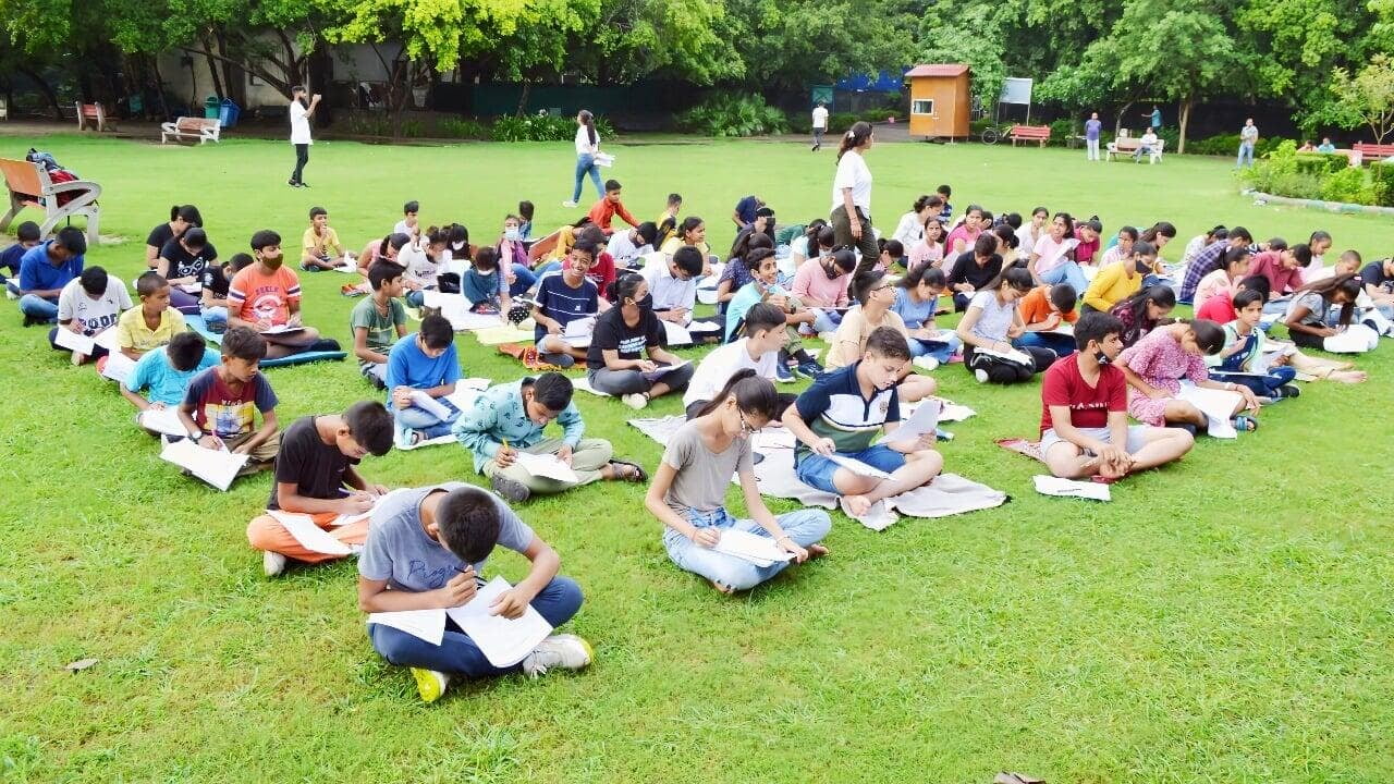 Group of students sitting outdoors on a grassy field during a chemistry learning activity. They’re writing in notebooks while teachers guide them, possibly working on exercises about chemical reactions, balancing equations, or preparing for a hands-on experiment. The open-air setting makes it feel like a relaxed science workshop where students can think, discuss, and learn chemistry in a more engaging way than a regular classroom.